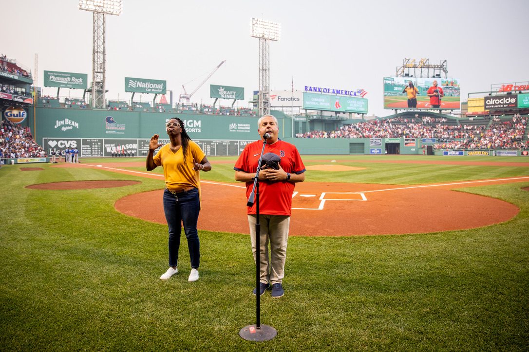 Standing on the grass at Fenway Park in a Red Sox jersey and kippah, cap held over his heart, singing into a microphone, with an ASL interpreter beside him and the Green Monster behind.