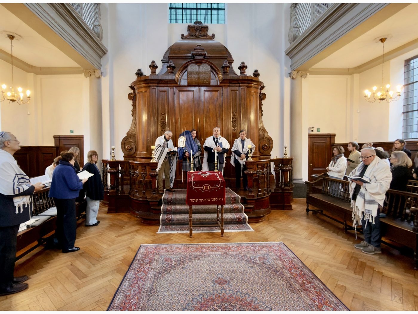 Interior of Beth Jehoeda during a service: a tall carved walnut ark at the centre, a reading desk draped in red before it, a congregation in prayer shawls standing on either side, and a Persian carpet on the parquet floor.