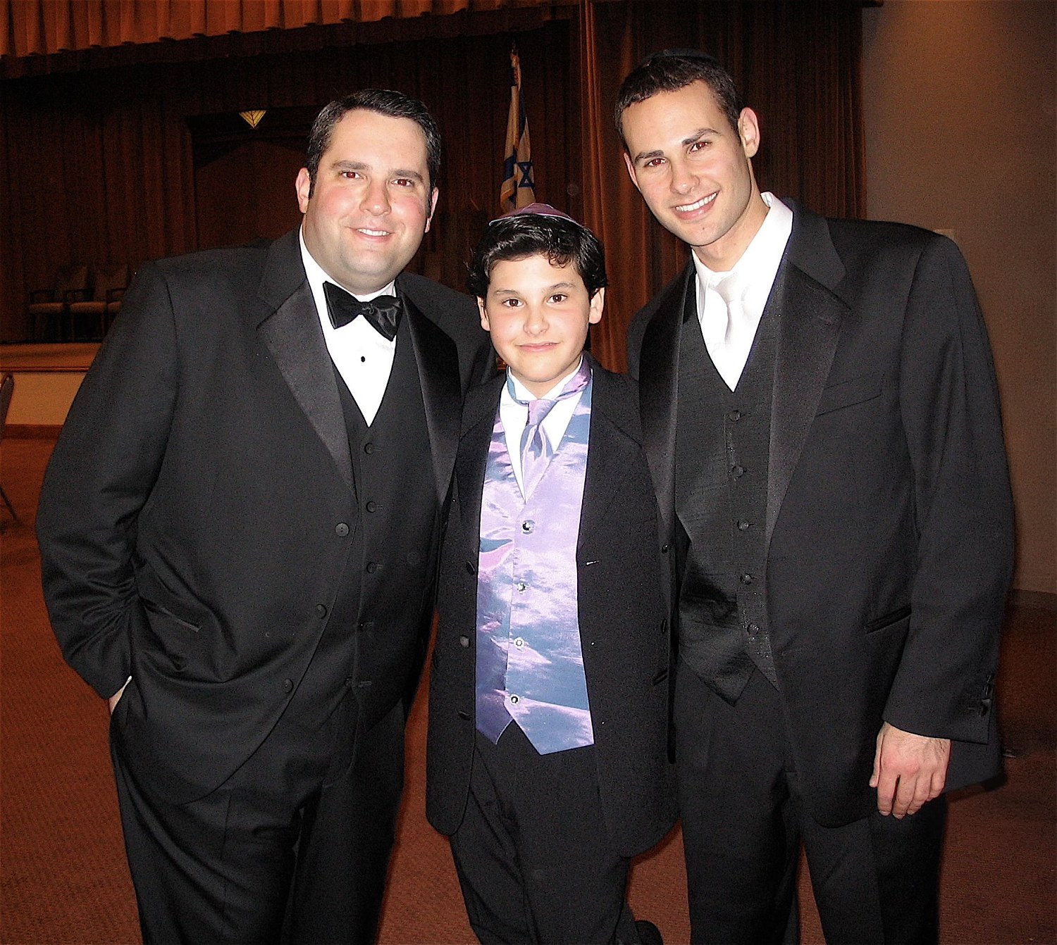 Three in black tie after the concert: Gideon Zelermyer at left, Jakie Young in the centre and Azi Schwartz at right, before the curtained bimah with an Israeli flag behind.