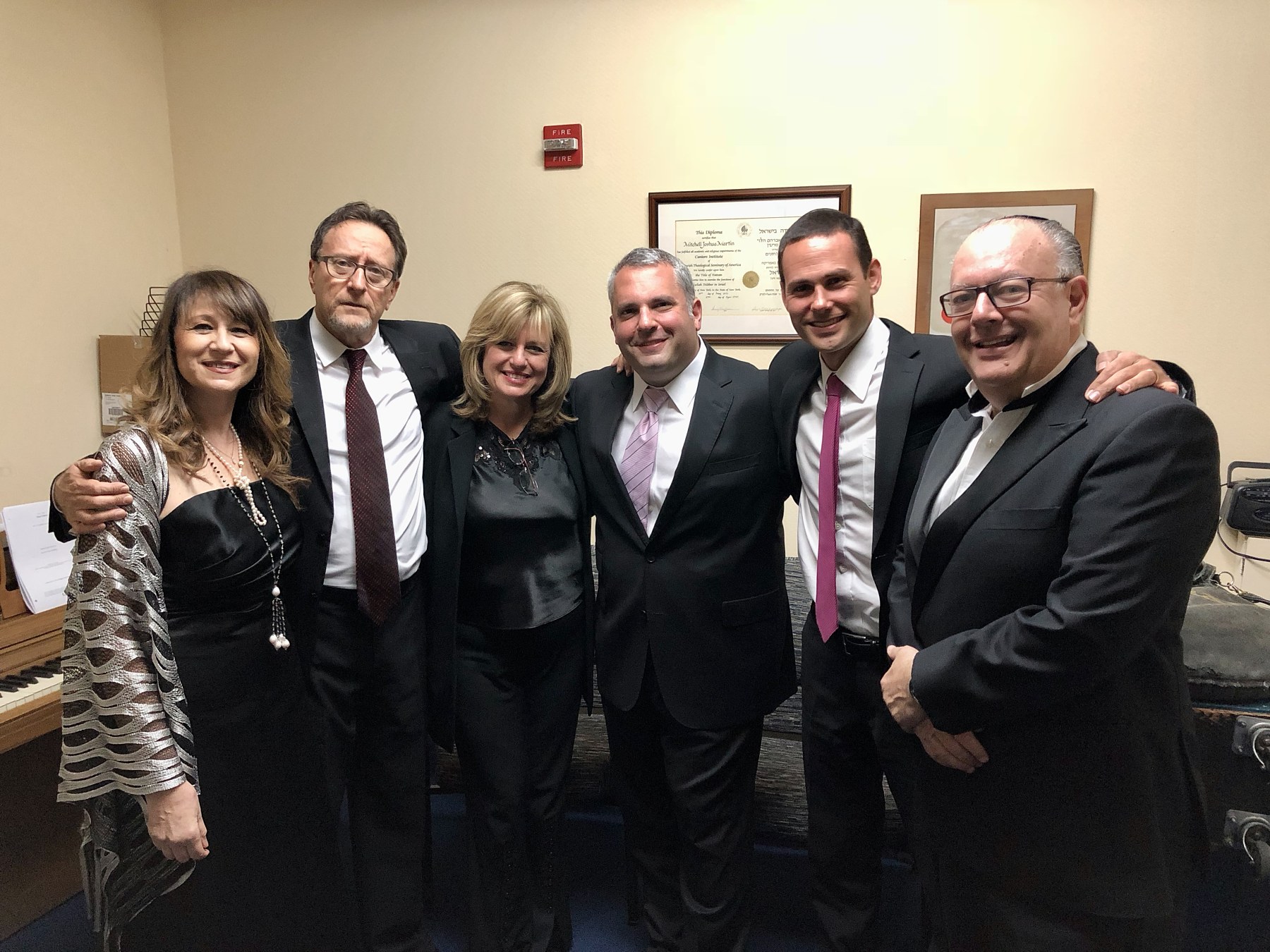 Six performers backstage in a rehearsal room, arms round one another, a framed cantorial diploma on the wall behind and a piano at the edge of frame.