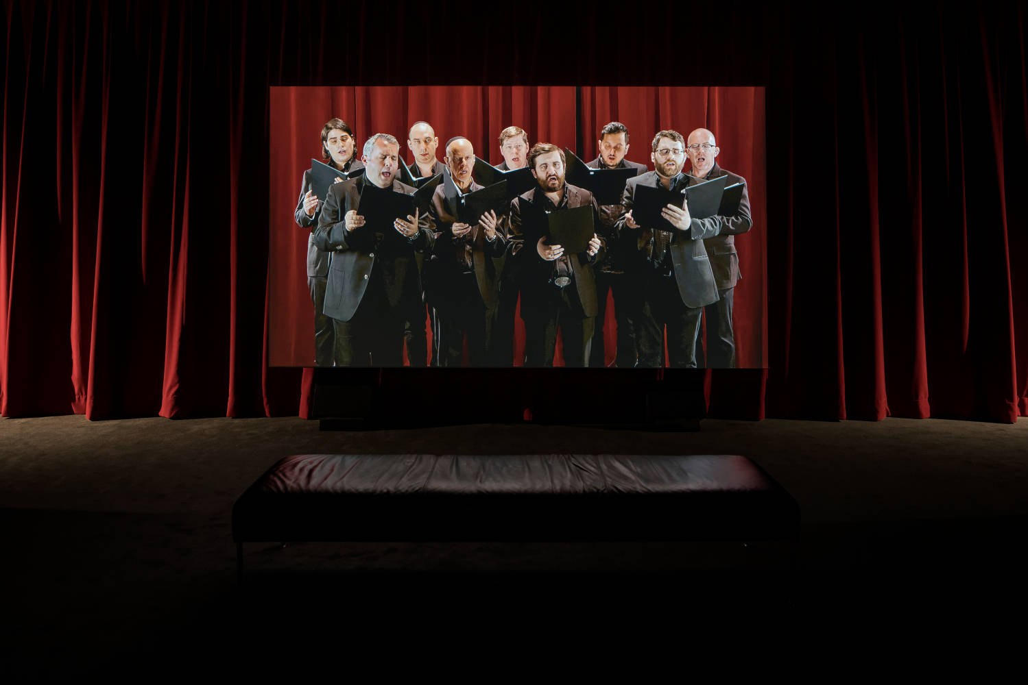 A large screen suspended in a darkened gallery hung with red curtains, showing nine men of the Shaar Hashomayim choir in dark suits singing from black folders; a bench sits empty in front of it.