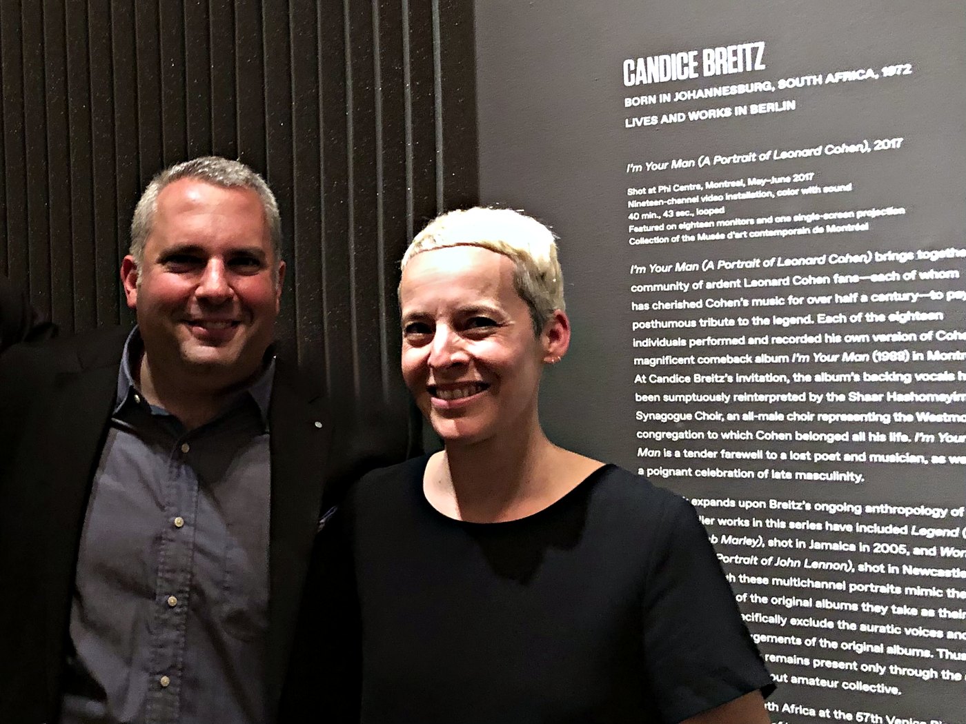 Gideon Zelermyer and Candice Breitz standing together, smiling, beside the exhibition wall text for I&rsquo;m Your Man (A Portrait of Leonard Cohen), which names the Shaar Hashomayim Synagogue Choir.