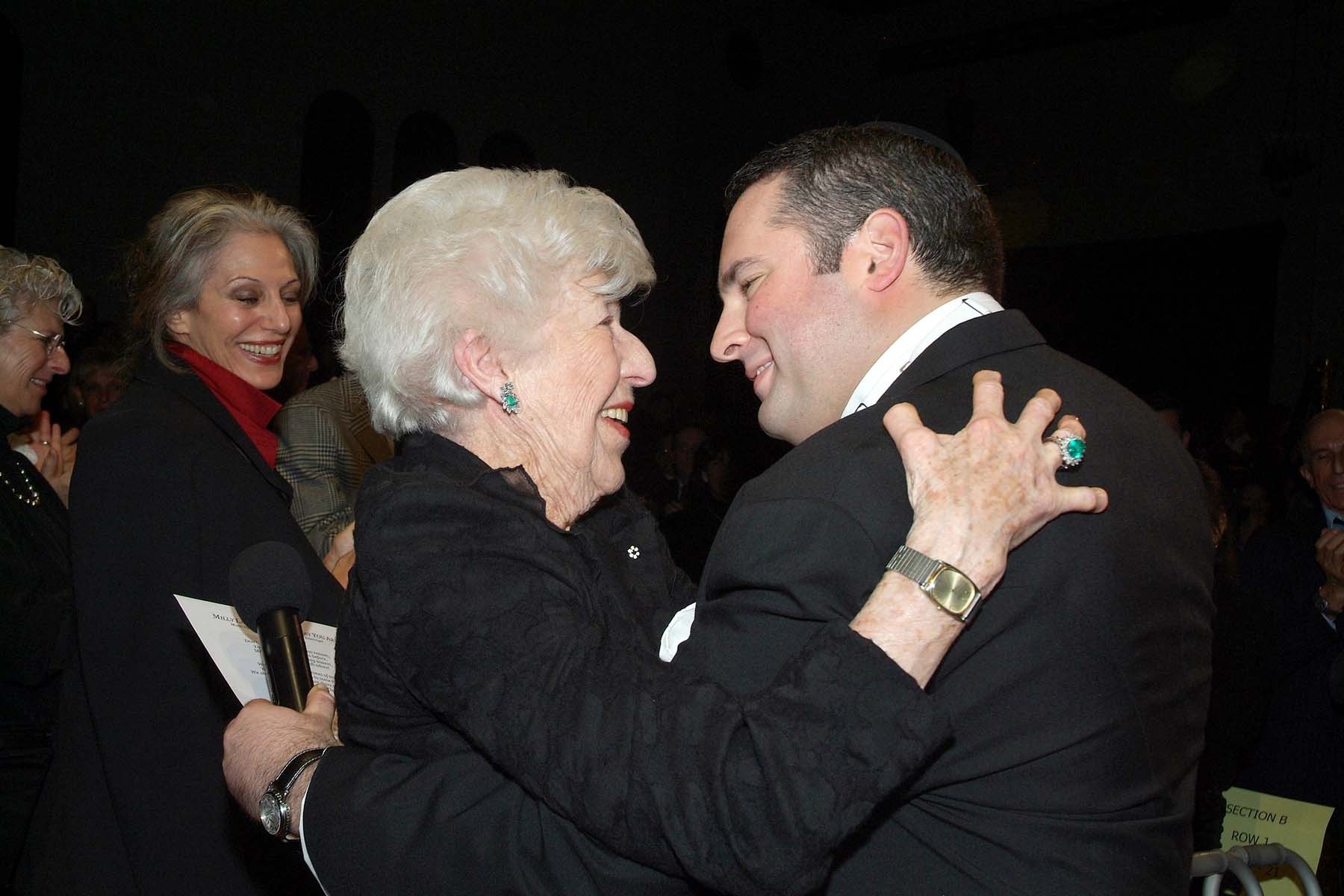 Milly Lande, wearing the Order of Canada and holding a microphone and a printed page, embracing Zelermyer with one hand on his shoulder, both smiling.