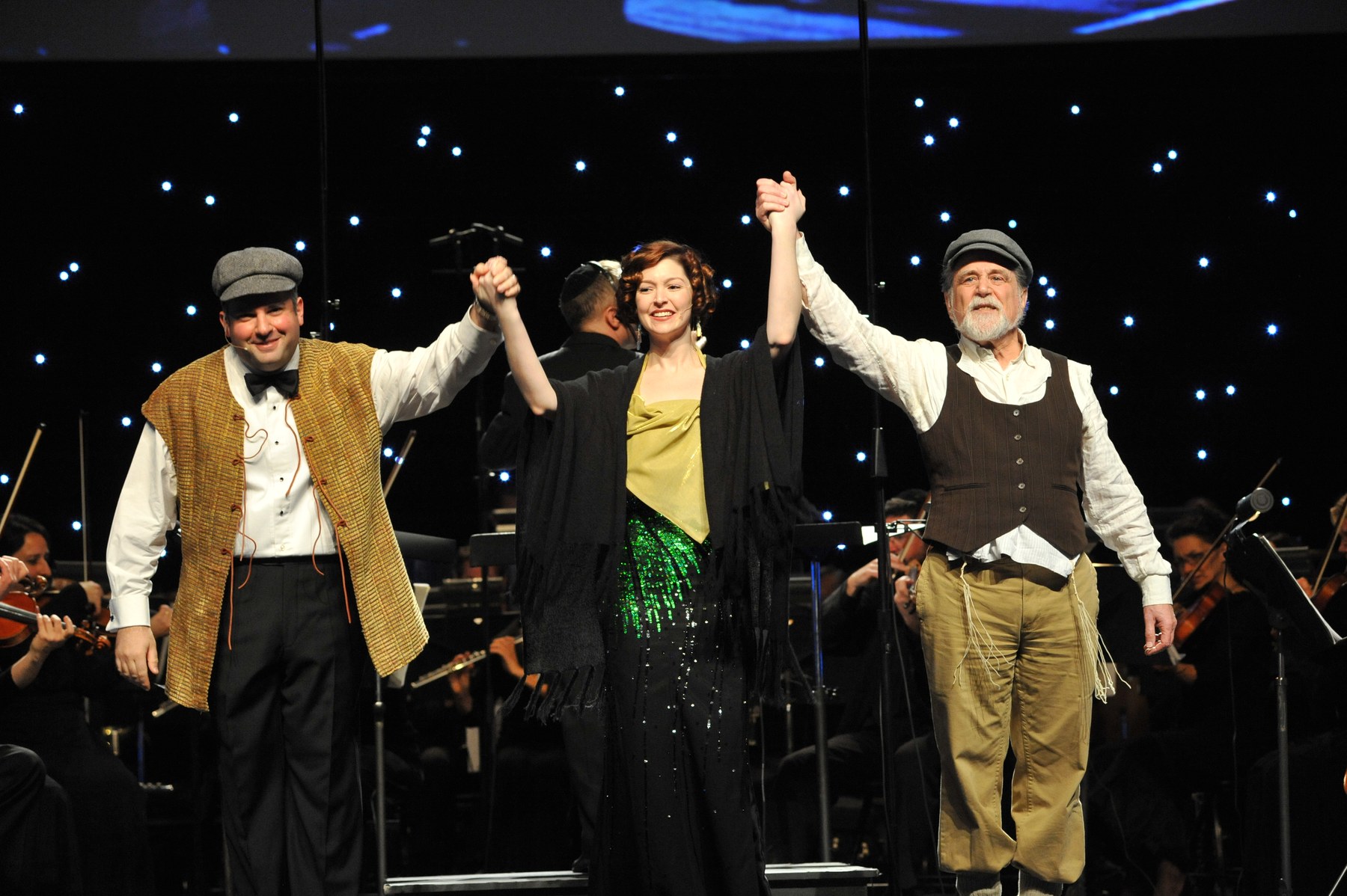 Zelermyer in flat cap and waistcoat, Leah Edwards in a green sequined gown and Eddie Aronson in cap, waistcoat and tzitzit, bowing hand in hand beneath a starcloth.
