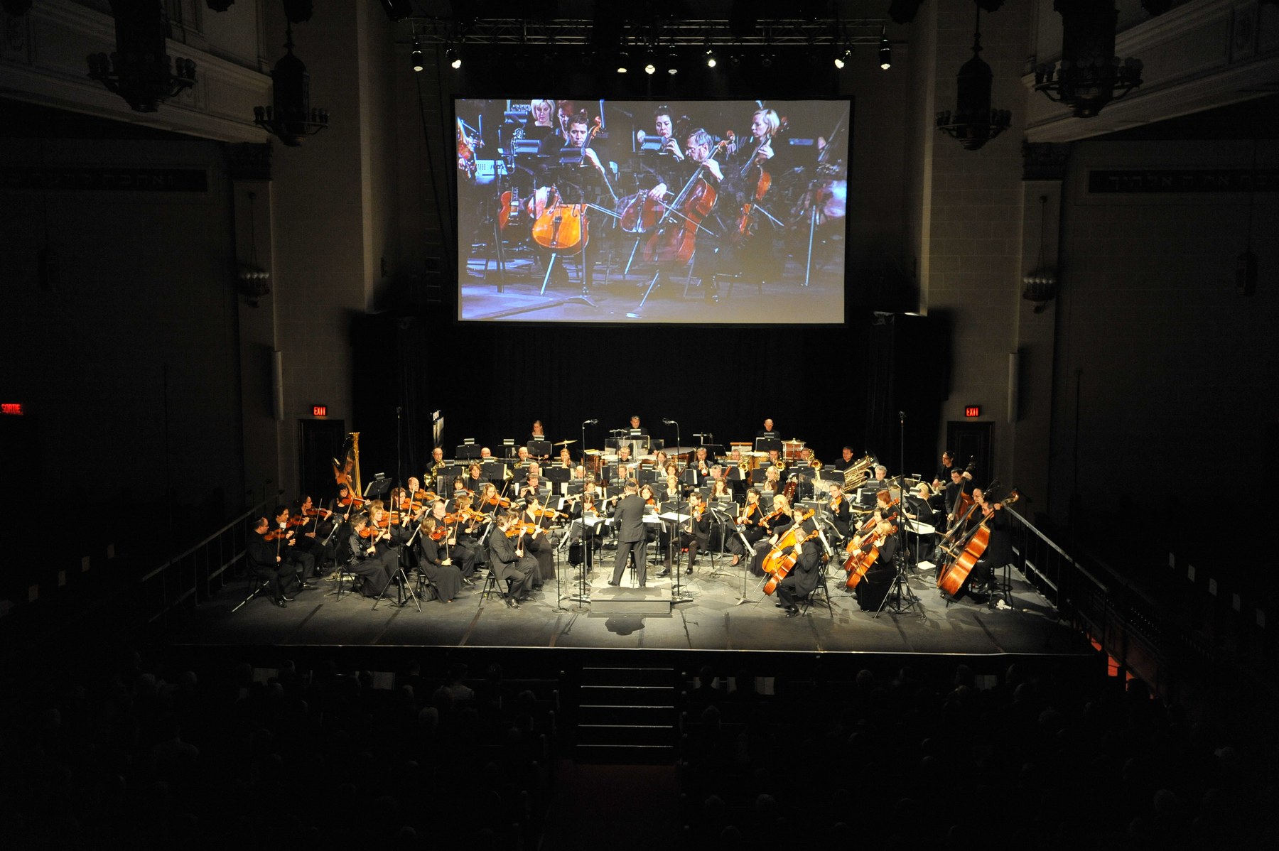 The sanctuary in darkness, a full symphony orchestra on a stage built out over the bimah, a wide screen above them showing the cello section in close-up.