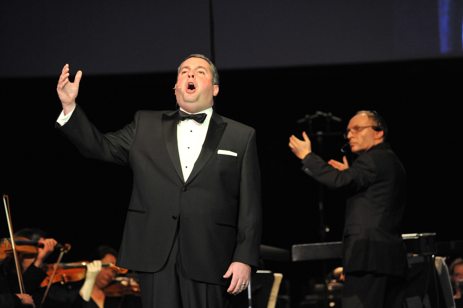 Zelermyer singing in black tie with one arm raised, Stephen Glass conducting behind him in a headset microphone.