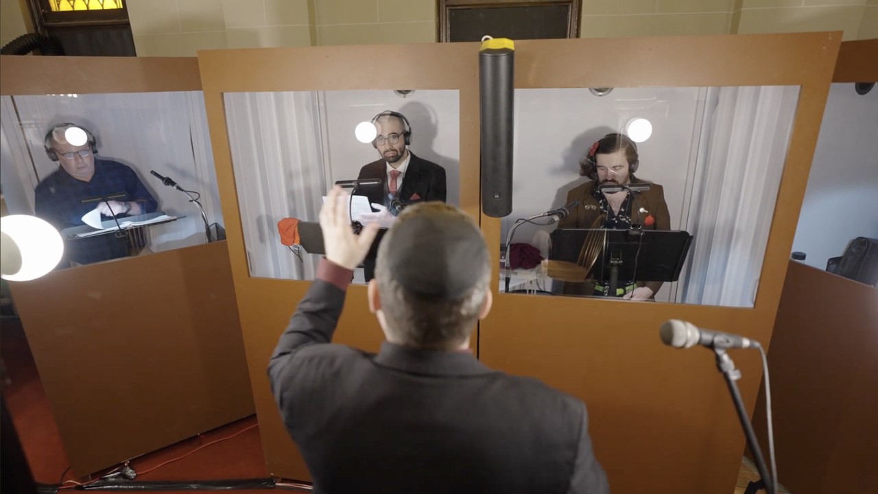 Three members of the choir singing in separate plywood booths with clear windows, each in headphones at his own microphone and stand, the conductor in the foreground with his hand raised, seen from behind.