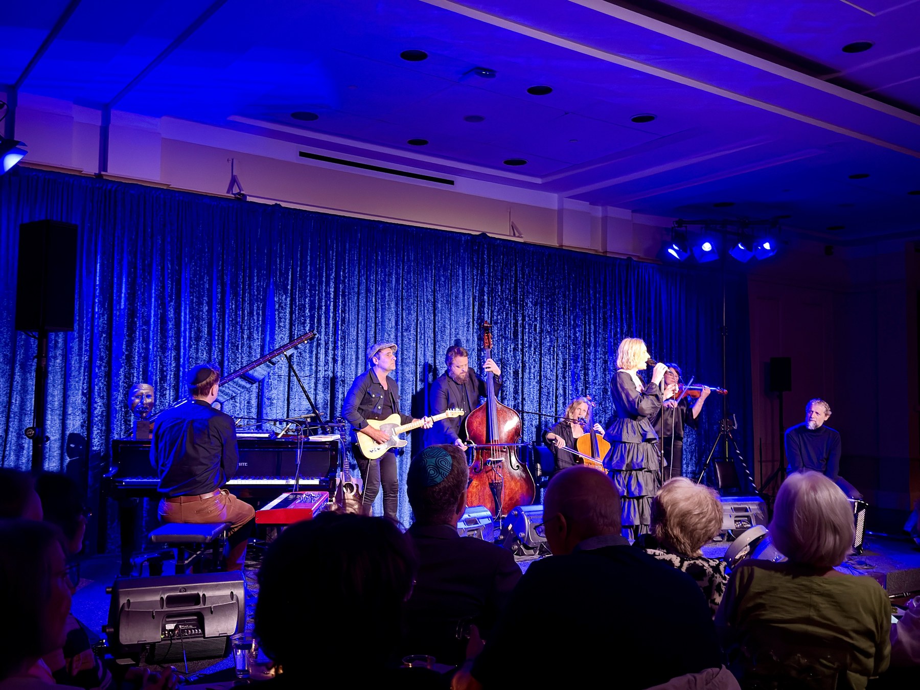 A band on a stage lit deep blue before a velvet curtain: pianist at a grand piano, guitarist in a flat cap, double bass, cello, violin, and Louisa Lyne singing into a hand-held microphone, with a full audience in the foreground.