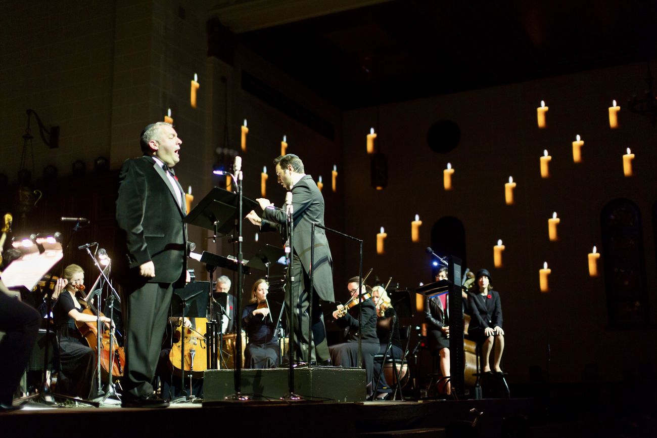 Zelermyer singing at a music stand beside a conductor, the sanctuary wall behind them covered in projected candle flames.