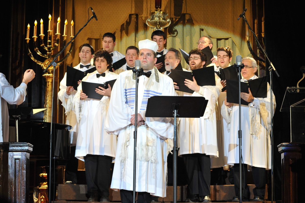 Zelermyer in white canonical robes and cantor's hat at a microphone, the male choir behind him in white robes and tallitot, a large candelabrum at left.