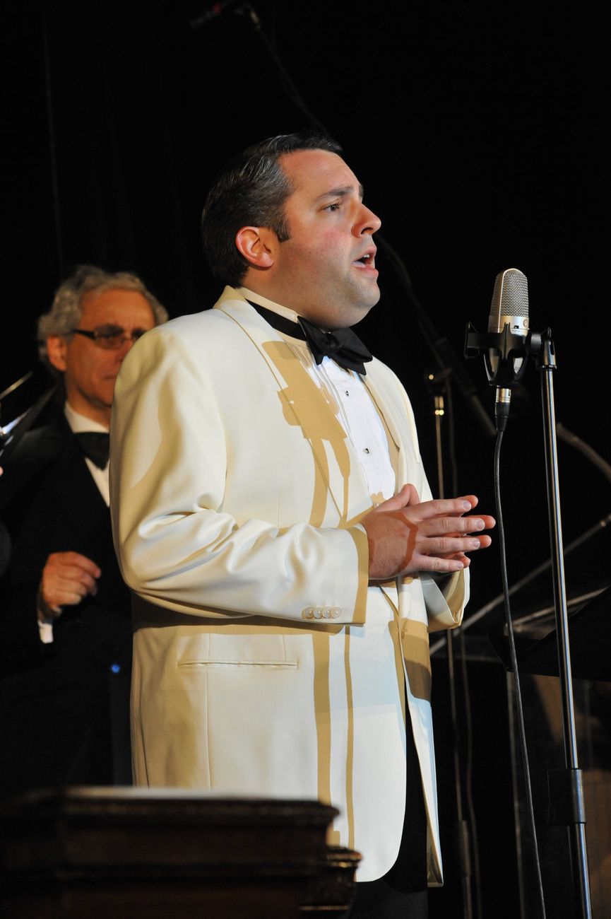Singing at a ribbon microphone in a cream dinner jacket and black bow tie, a pianist behind.