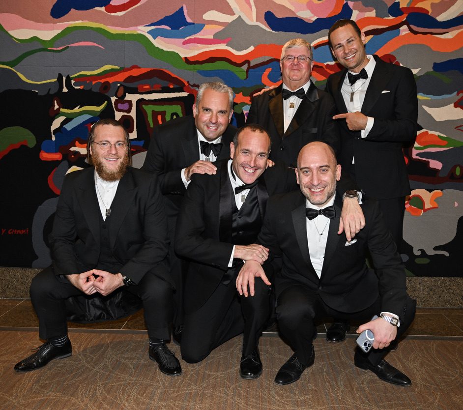 Six men in black tie posed together after the concert, several kneeling, one pointing at another, in front of a large abstract tapestry.