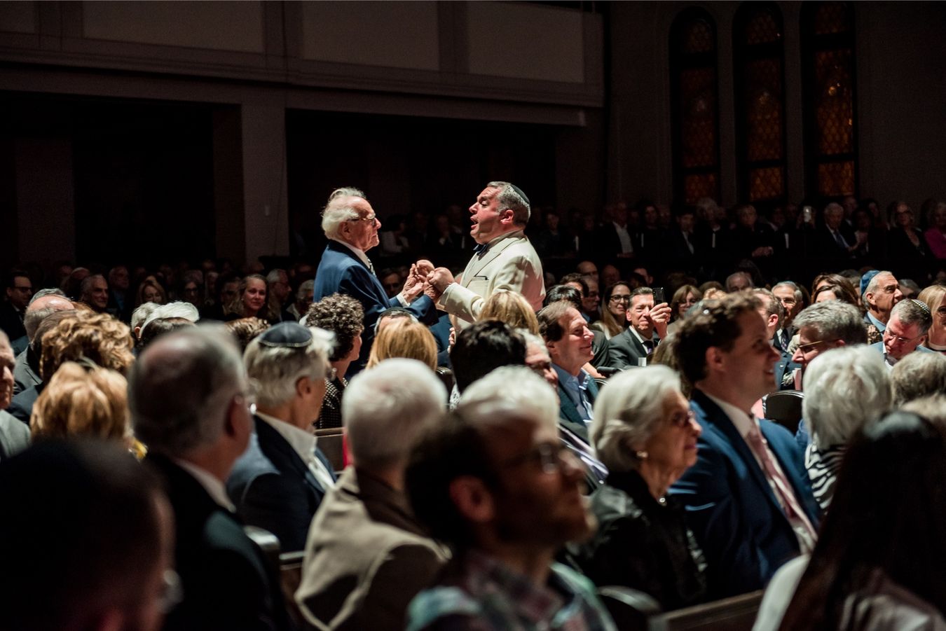 Zelermyer singing in a white dinner jacket from the middle of a packed audience, holding an older man&rsquo;s hand.