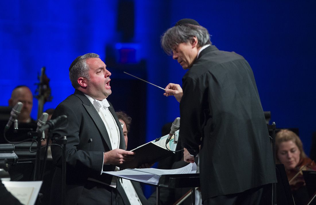 Gideon Zelermyer singing from a score, facing Kent Nagano, who conducts with a baton raised.