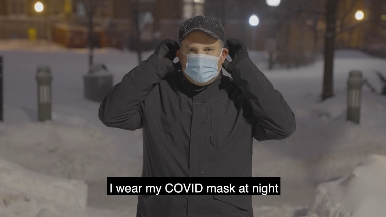A man in a dark winter coat, cap and gloves fitting a surgical mask over his ears on a snowbanked Montreal street at night, streetlamps behind him, a subtitle reading &ldquo;I wear my COVID mask at night&rdquo;.
