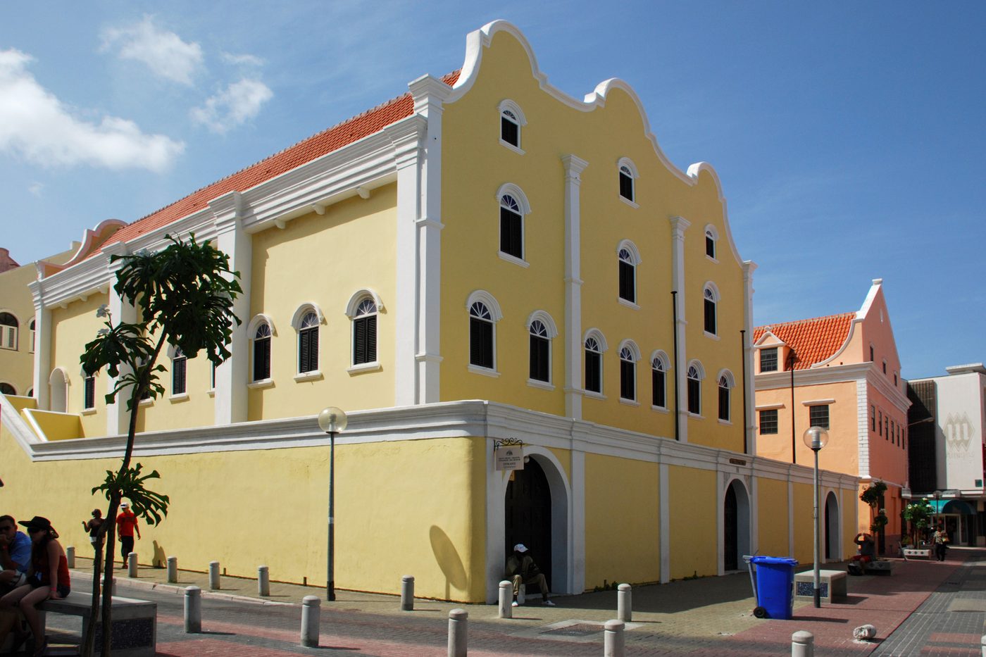 The Mikvé Israel-Emanuel synagogue in Willemstad: a yellow building with white trim, a scrolled Dutch gable and a red tile roof, seen from the corner of the street under a blue sky.