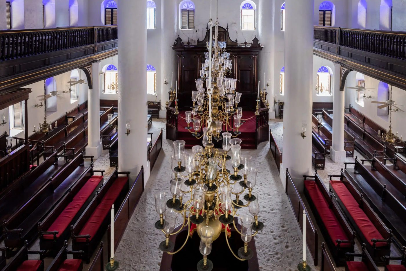 The interior seen from the gallery: dark mahogany pews with red cushions on either side of a white sand floor, brass chandeliers hanging the length of the room, and the ark at the far end.