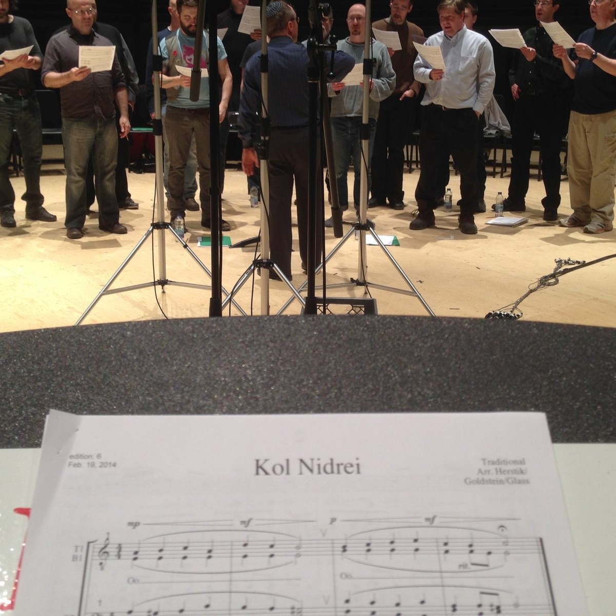 The Shaar Hashomayim choir recording in a hall, seen past a music stand in the foreground holding the Kol Nidrei score, with microphone stands between.