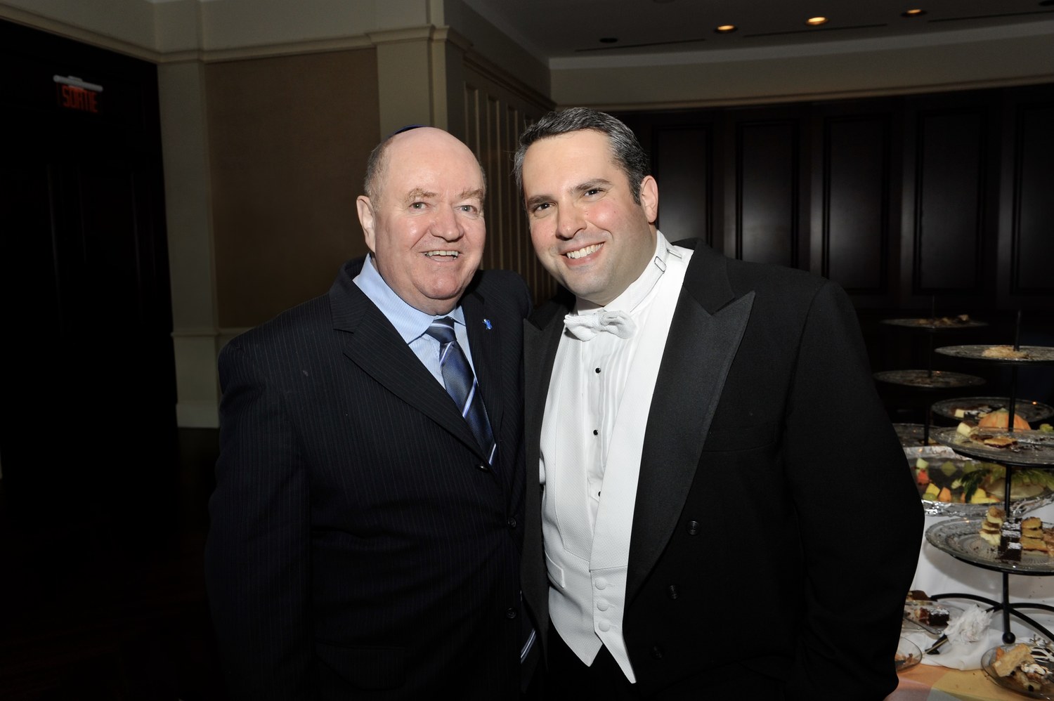 Father John Walsh in a dark suit and kippah with his arm around Gideon Zelermyer in white tie, both laughing, a tiered dessert stand behind them.