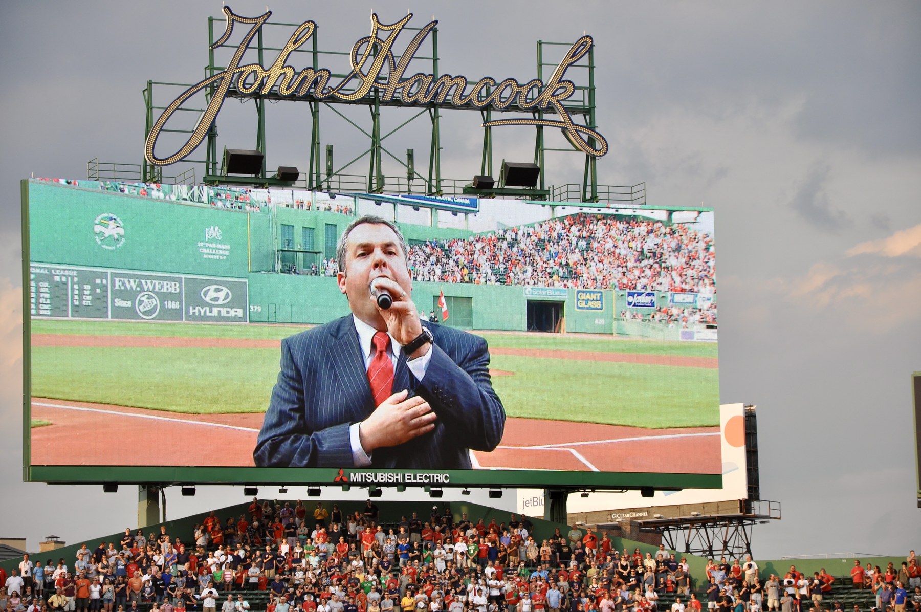 The Fenway Park scoreboard beneath the John Hancock sign, showing Zelermyer in a suit and red tie singing with his hand over his heart, the standing crowd in the bleachers below.