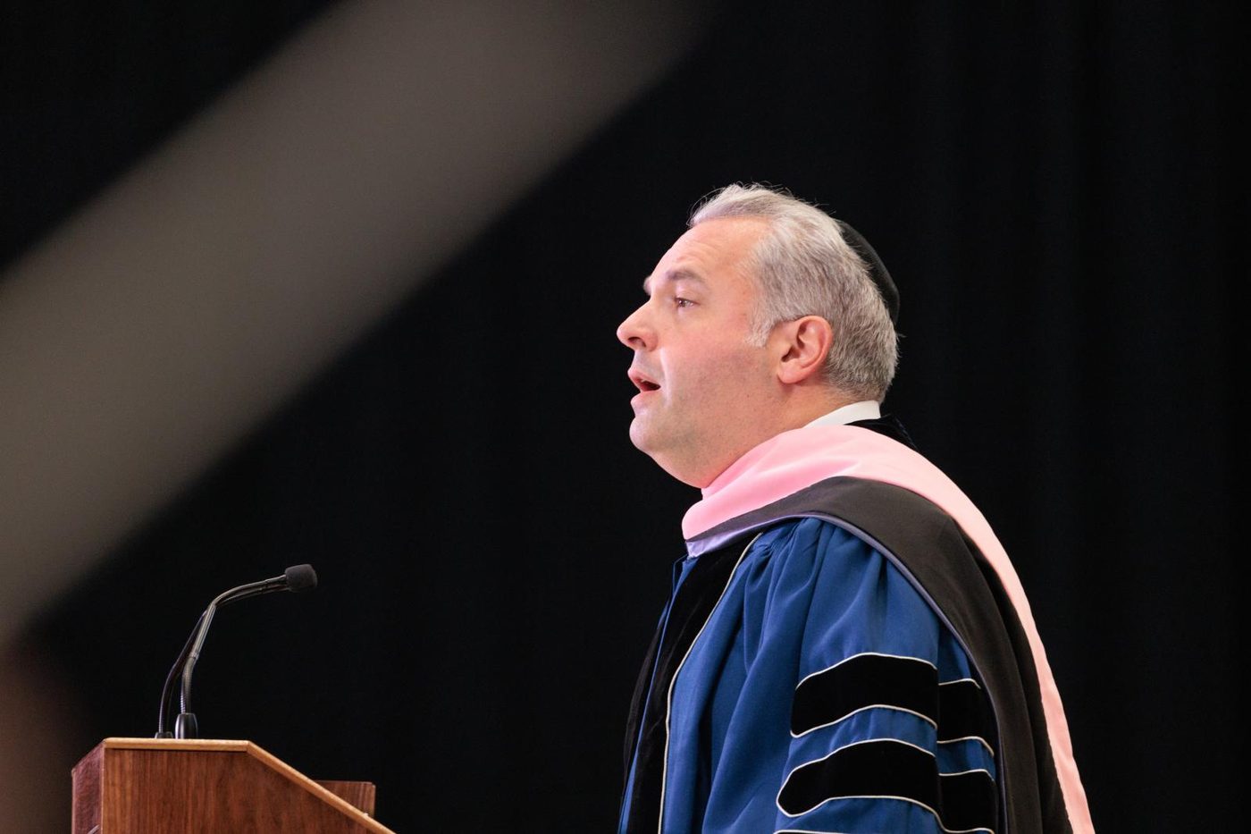 Gideon Zelermyer in academic robes and kippah, speaking at a lectern in profile.