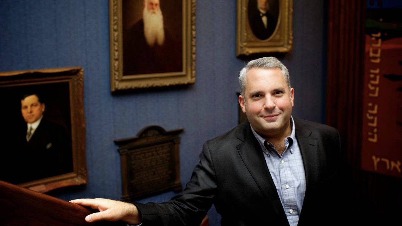 Zelermyer in a dark jacket, leaning on a wooden rail and smiling, before a blue wall hung with gilt-framed portraits of two men in nineteenth-century dress, with a red Torah mantle lettered in gold at the right.
