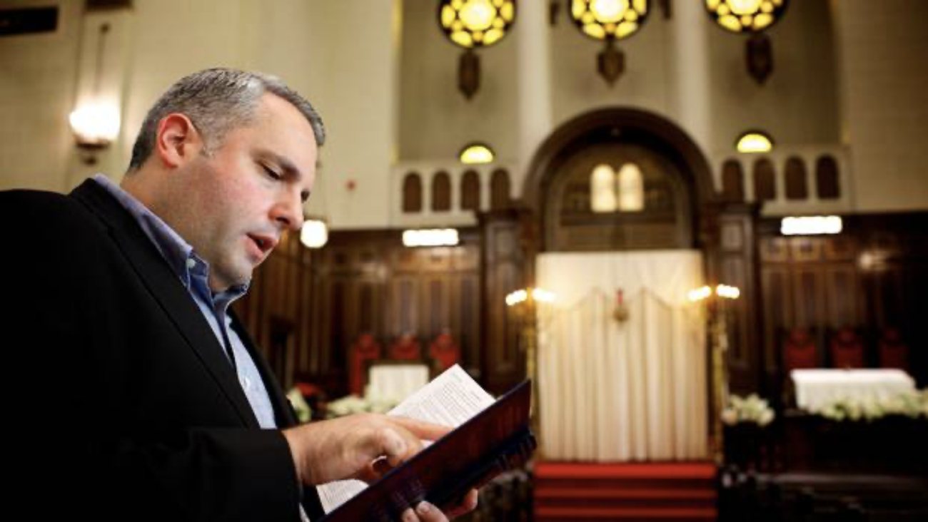 Gideon Zelermyer in a dark jacket and open-necked blue shirt, singing from a small prayer book held in both hands, in the sanctuary of Shaar Hashomayim: the ark curtained in white behind him, rose windows lit above, dark carved wood and red seats beyond.