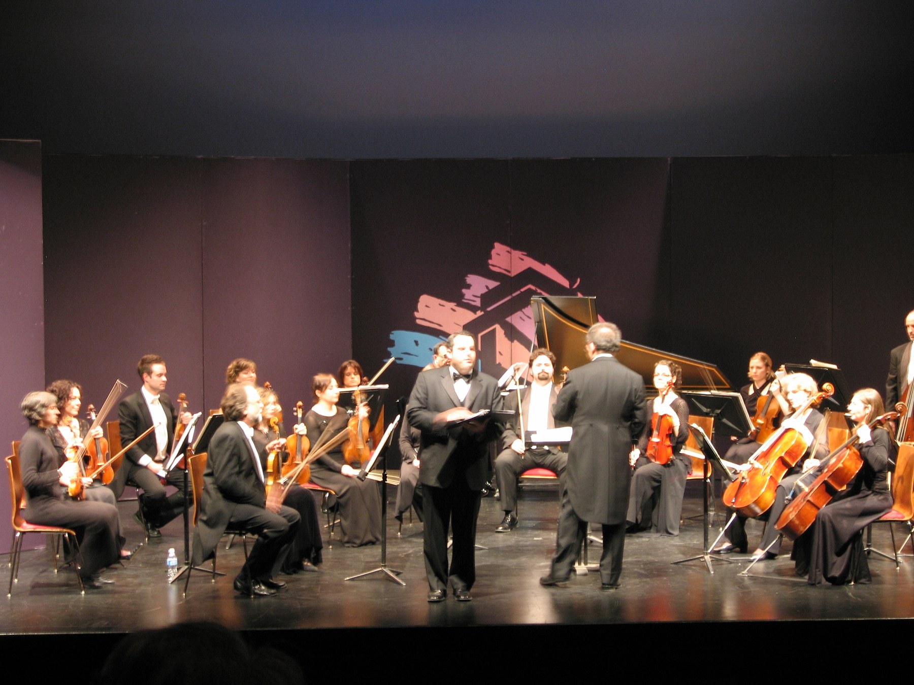 On a theatre stage, Zelermyer in white tie stands score in hand before the seated strings of the McGill Chamber Orchestra, the conductor's back to the camera and a harpsichord behind.
