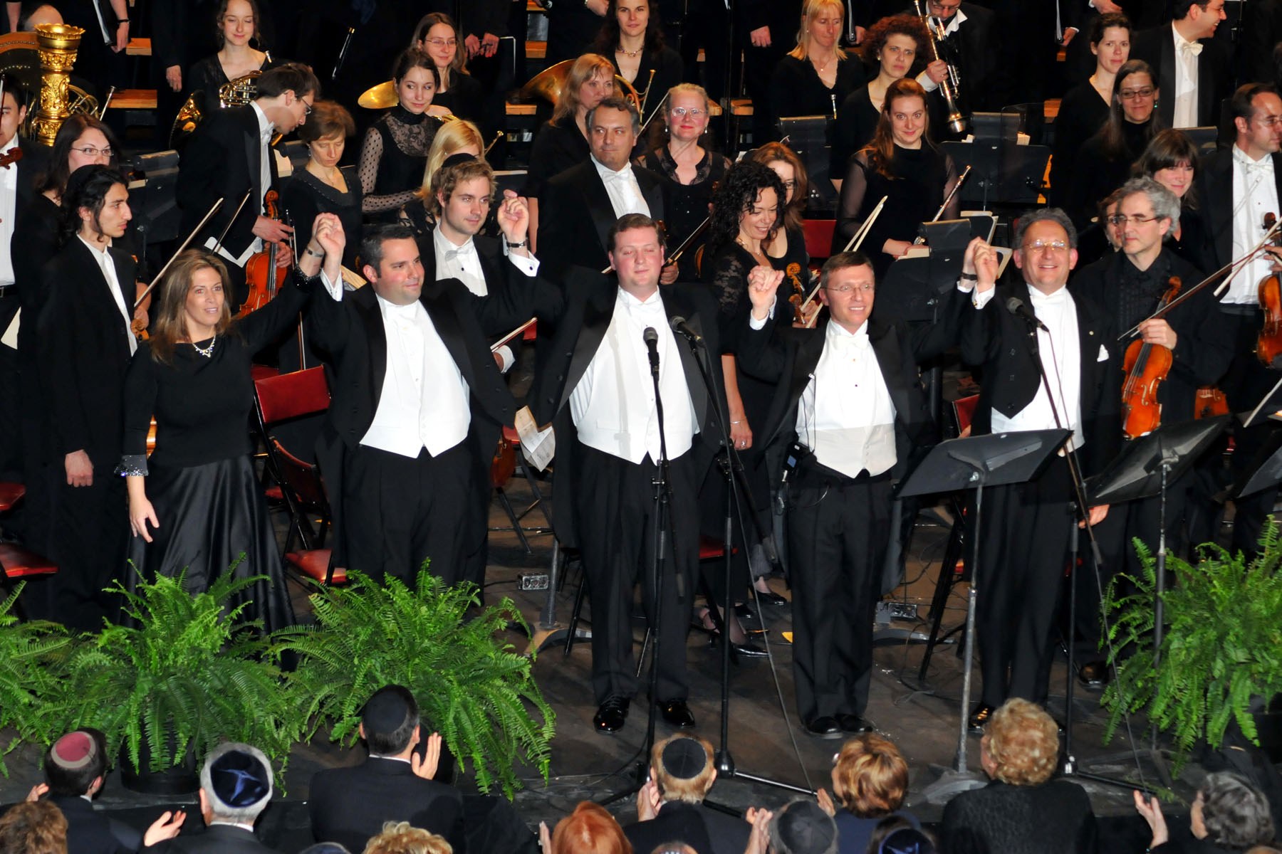 Five performers taking a bow with hands joined and raised, the orchestra applauding behind them.