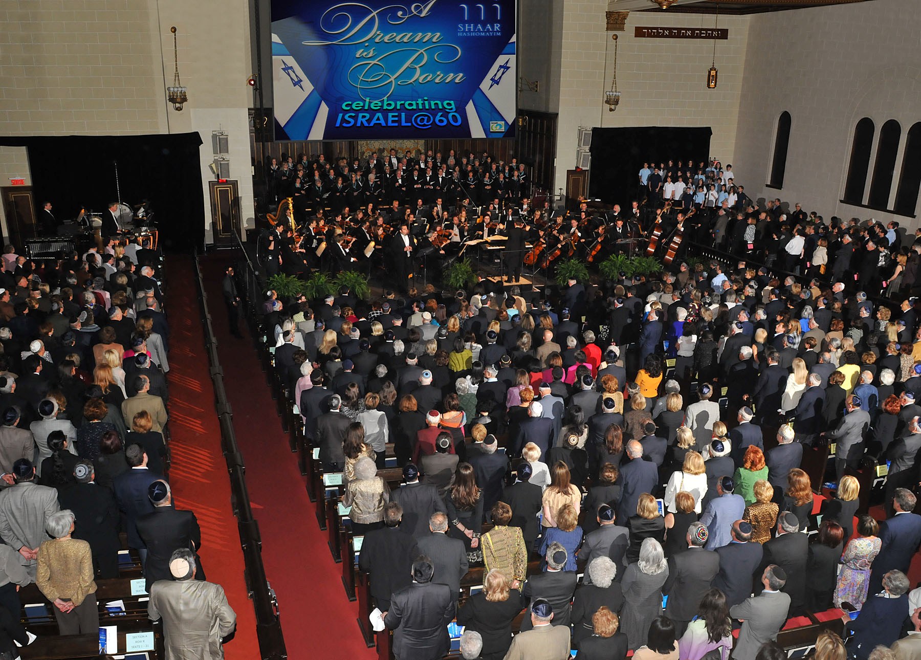 The sanctuary from the balcony, the congregation standing, orchestra and two choirs on the bimah beneath a screen reading A Dream is Born, celebrating Israel@60.