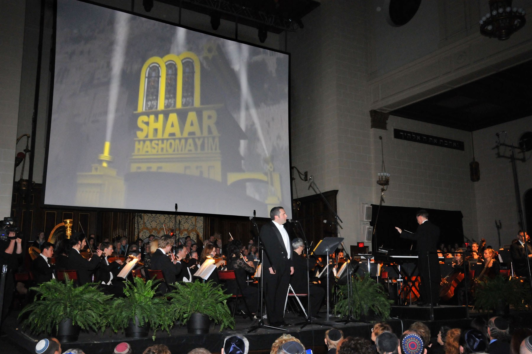 Zelermyer in white tie singing at a microphone in front of the orchestra, the conductor at right, the screen above showing the Shaar Hashomayim building in yellow.