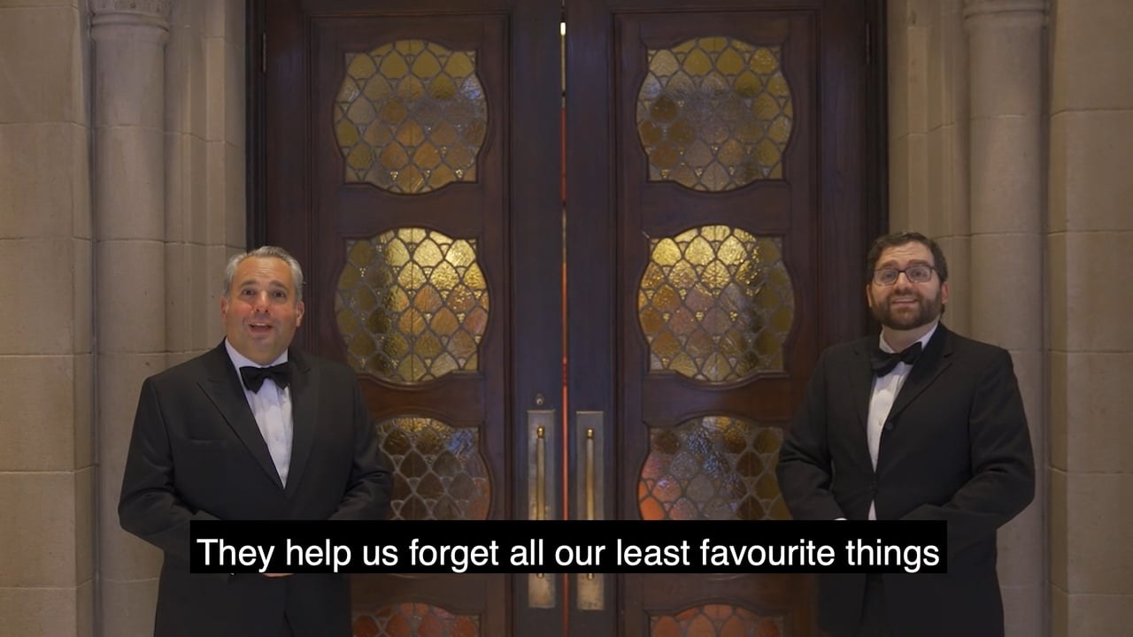 Two men in black tie singing side by side before tall carved wooden doors set with leaded amber glass, a subtitle across the foot of the frame reading &ldquo;They help us forget all our least favourite things&rdquo;.