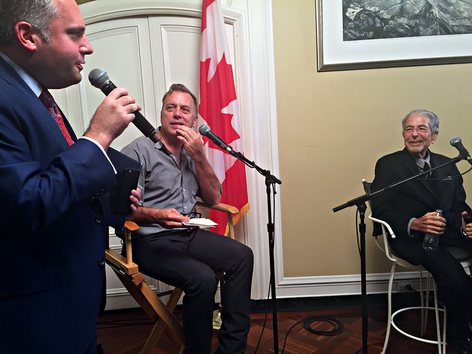 Gideon Zelermyer speaking into a microphone at the album launch, with Leonard Cohen seated at right, smiling.
