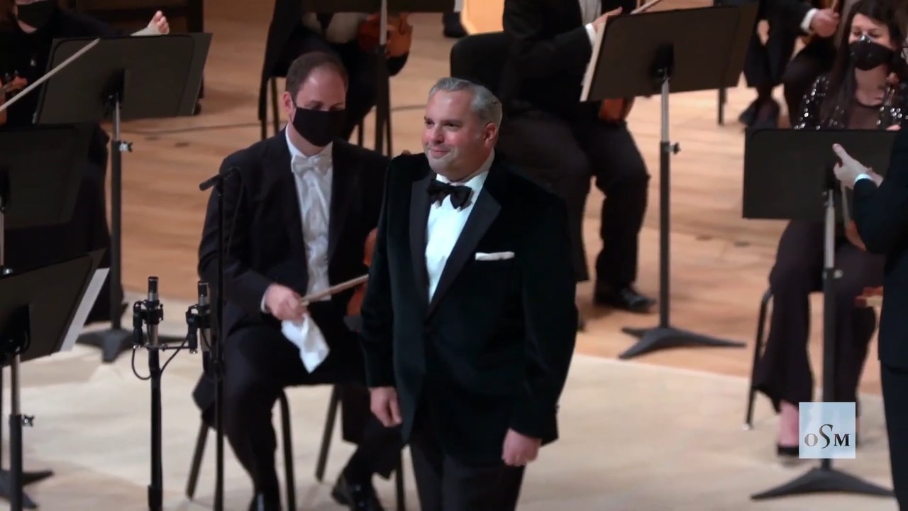 Zelermyer in black tie walking forward on the Maison symphonique stage, the masked players of the Orchestre symphonique de Montr&eacute;al seated behind him among microphone stands.