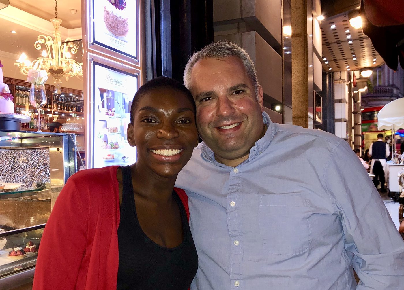 Gideon Zelermyer and Michaela Coel standing cheek to cheek outside a café at Piccadilly Circus, both smiling at the camera.