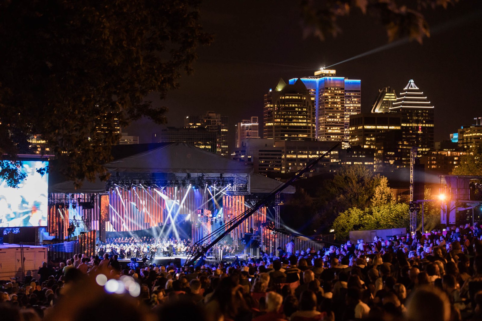 Night view over the crowd at the foot of Mount Royal: the lit stage with orchestra and choir, a screen at left, and the downtown Montreal skyline behind.