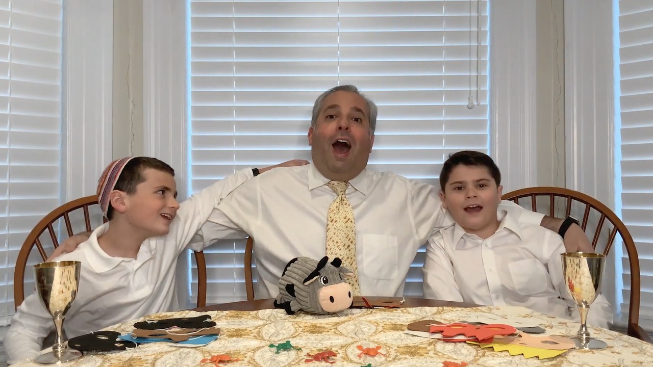 Zelermyer at the seder table in a matzah-patterned tie, an arm round each of his two sons, all three singing; goblets, plague masks and plastic frogs on the cloth in front of them.