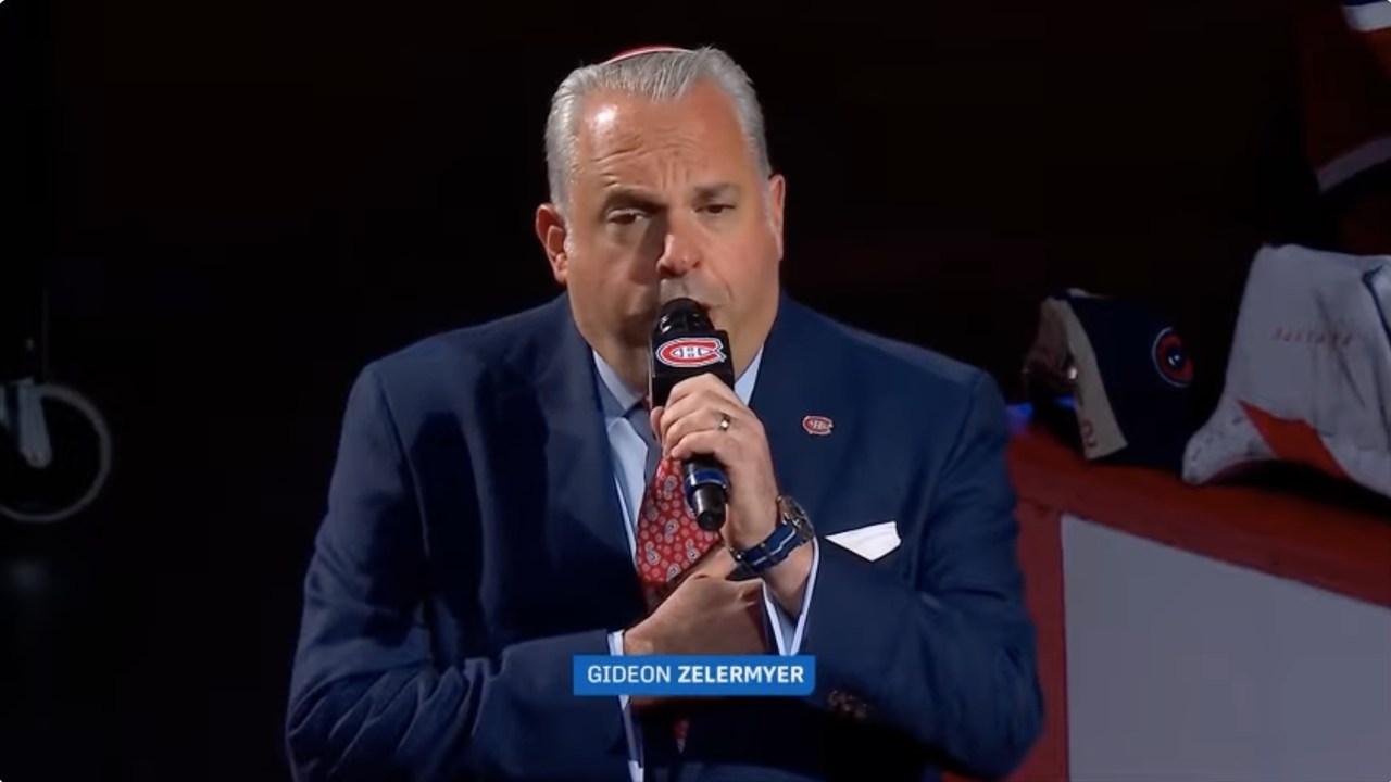 Zelermyer in a navy suit and kippah singing into a Canadiens microphone at the Bell Centre, a Canadiens tie clutched in his hand, a caption bar across the frame reading Gideon Zelermyer.
