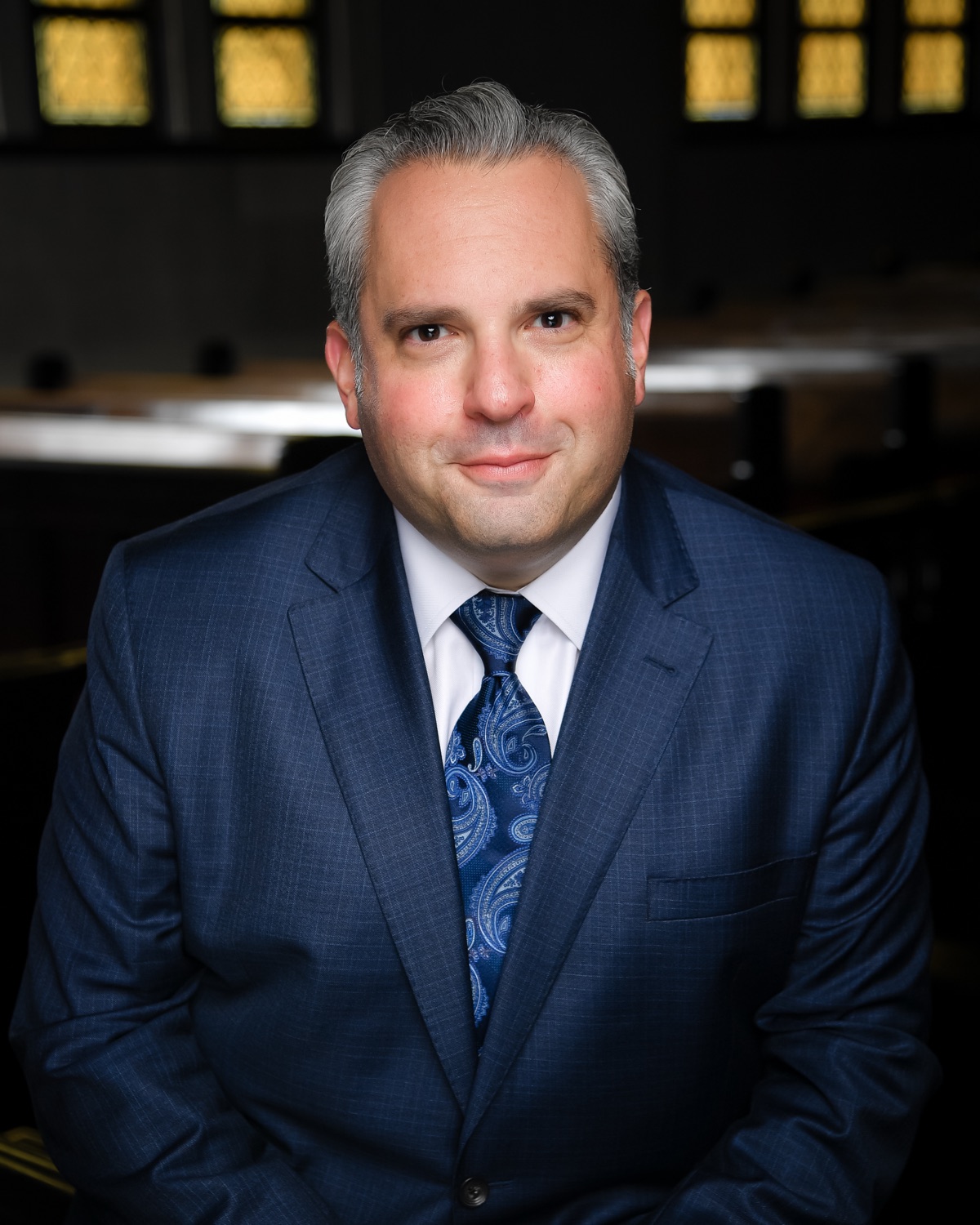 Portrait in a blue suit and tie, seated in the sanctuary.