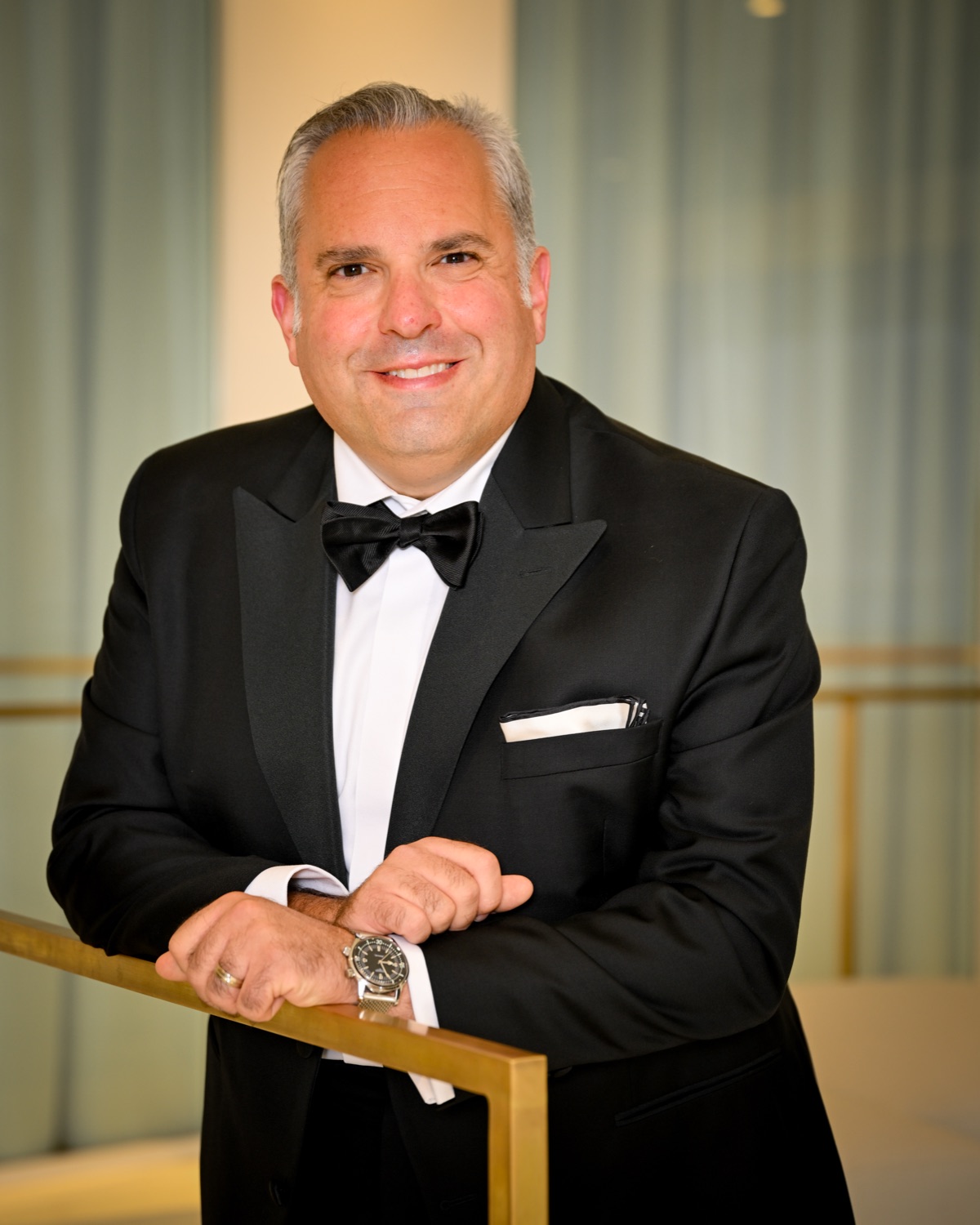 Portrait in black tie, leaning on a brass railing before a pale curtain.