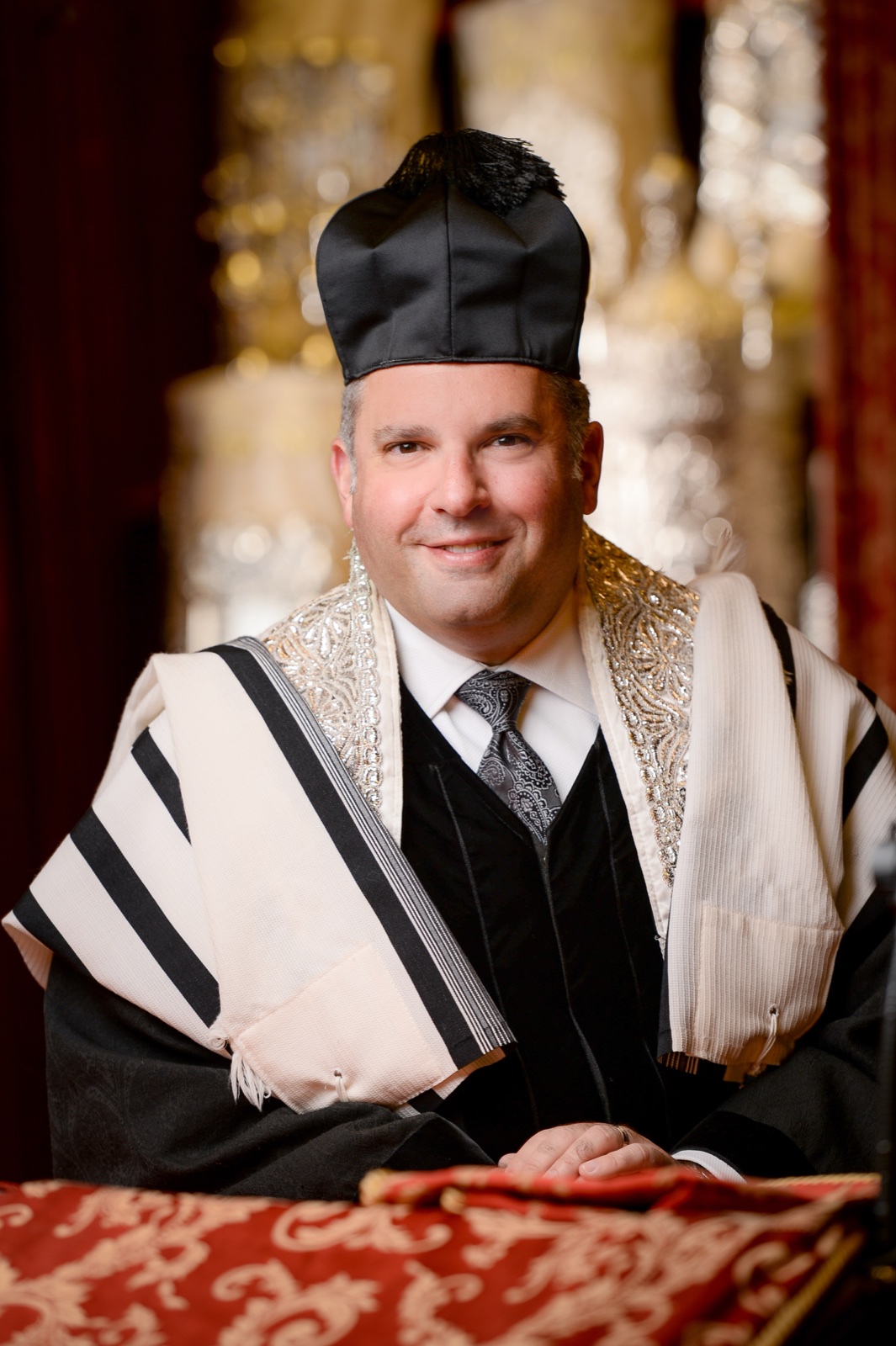 Portrait in cantor's robes, tallit and hat, seated at the reading desk before the ark.
