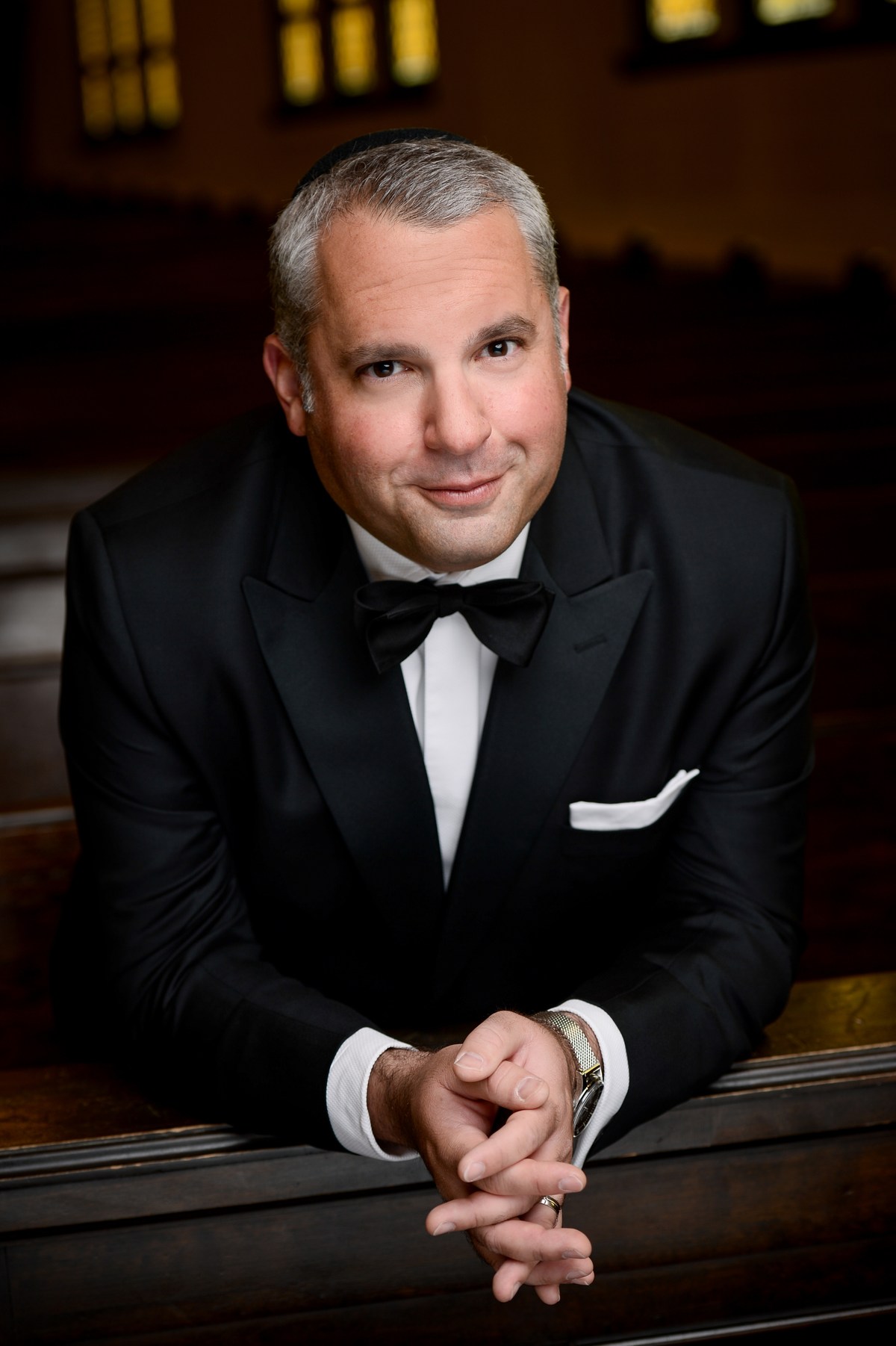Gideon Zelermyer in black tie and kippah, leaning on a pew rail in the sanctuary with his hands folded, looking to camera.