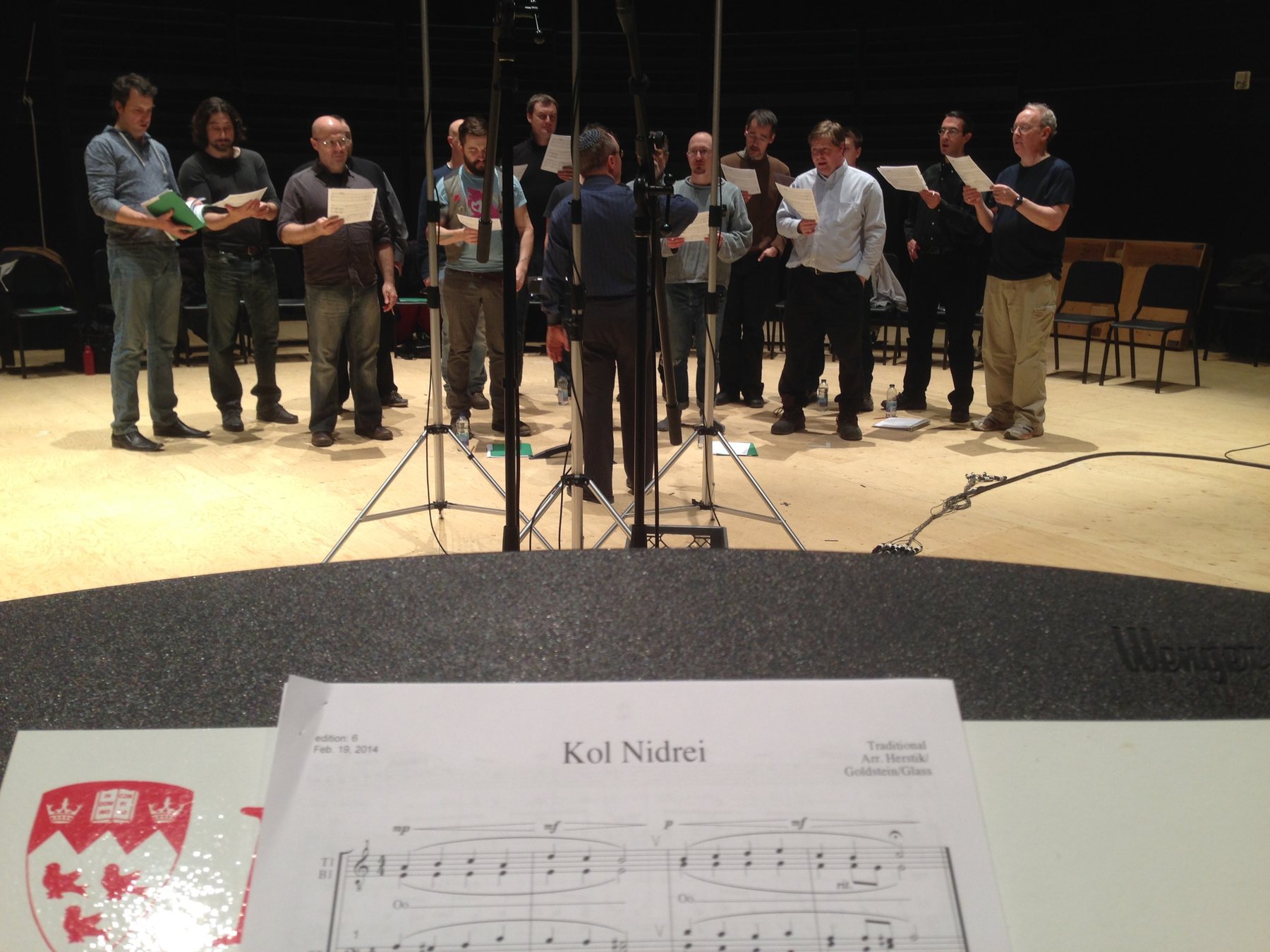 The men of the Shaar Hashomayim Synagogue Choir in shirtsleeves on a studio stage, scores in hand around a cluster of microphones, Stephen Glass conducting, his back to the camera; a copy of Kol Nidrei open on the music stand in the foreground.