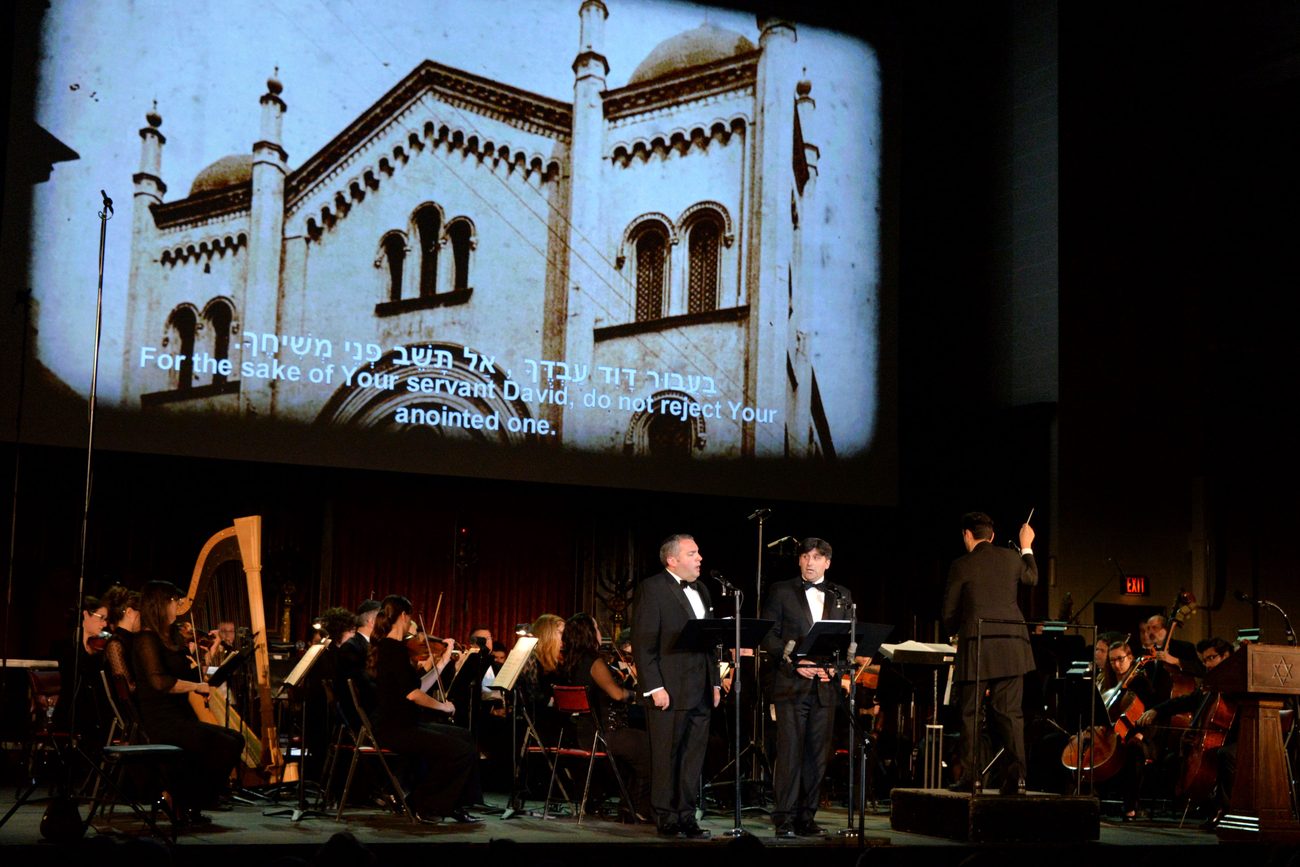 Gideon Zelermyer and Netanel Baram singing at music stands with a harp and orchestra behind them, a destroyed synagogue projected on the screen above with the Hebrew text and its English translation running across it.