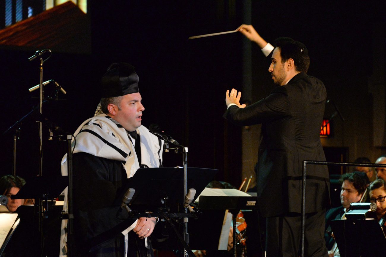 Gideon Zelermyer in canonical hat and tallit singing at a music stand, Roï Azoulay facing him with both hands raised.