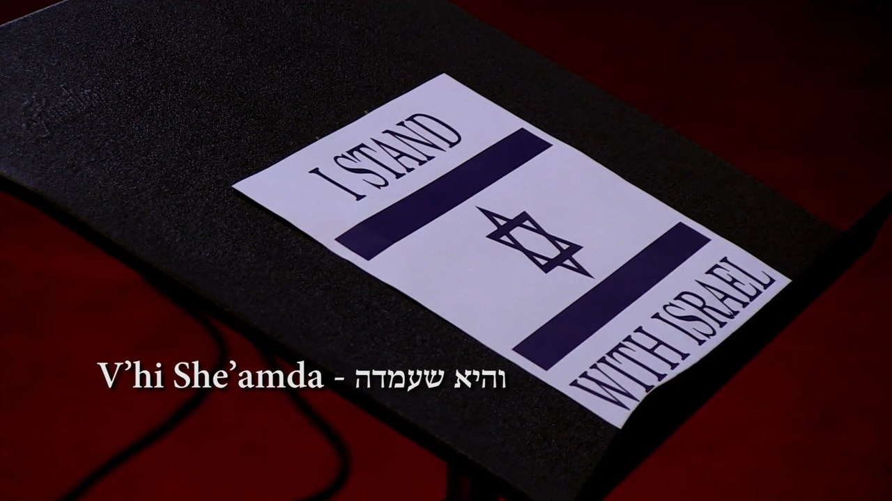 A printed card in the form of the Israeli flag reading &ldquo;I stand with Israel&rdquo;, lying on a black music stand above red carpet, with a title across the frame reading V&rsquo;hi She&rsquo;amda in Roman and Hebrew letters.