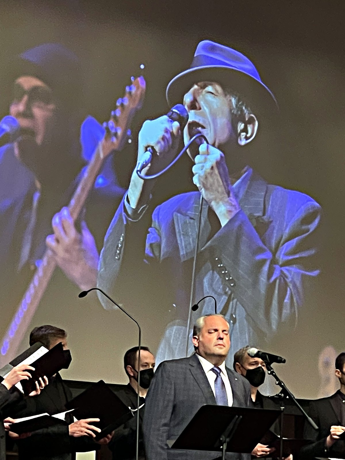Gideon Zelermyer singing at a music stand with the male choir behind him, Leonard Cohen in hat and suit projected on the screen above, singing into a microphone.