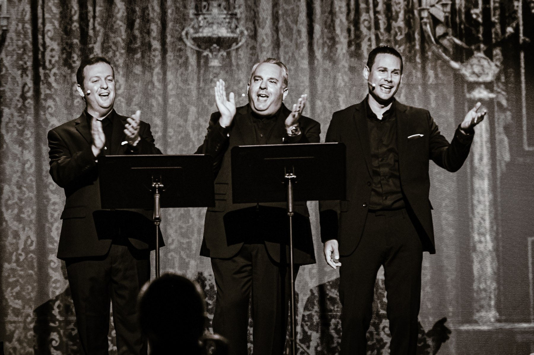 Three cantors in black, laughing and applauding at their music stands on a lit stage before a draped backdrop.