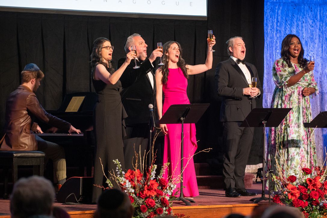 Five singers on stage raising champagne glasses mid-phrase, a pianist at a grand piano to the left, banked red roses along the front of the stage.