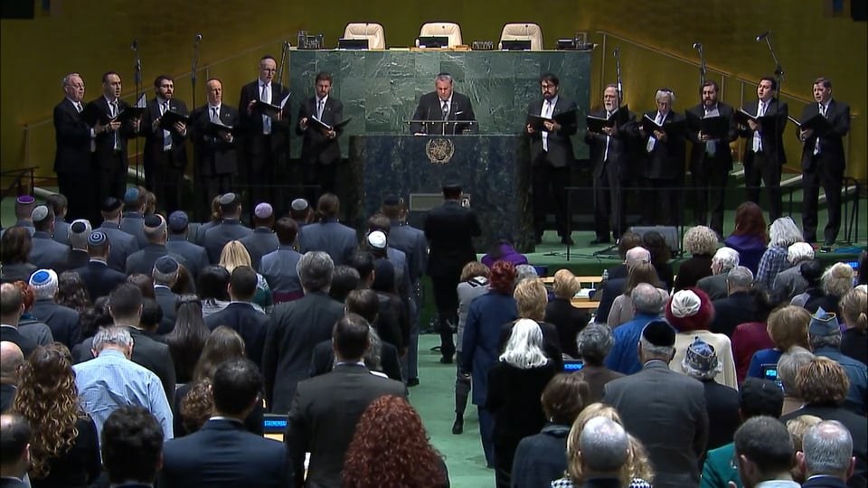 The Shaar Hashomayim choir in dark suits, standing in two ranks either side of the green marble rostrum of the United Nations General Assembly hall, folders open, with Zelermyer at the lectern between them and the seated assembly in the foreground.